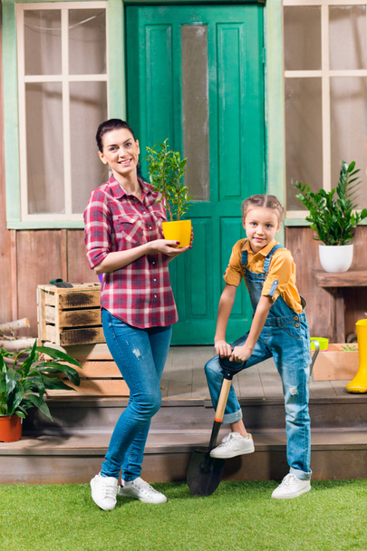 Smiling mother with potted plant and daughter with garden shovel standing on porch - Fotó, kép