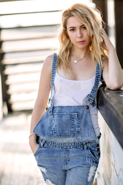 Young stylish blonde girl in denim overalls outdoors with natural daylight, a sunny day - Fotoğraf, Görsel
