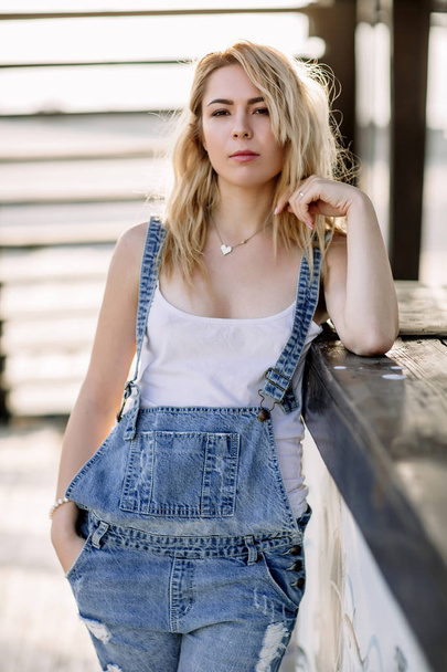 Young stylish blonde girl in denim overalls outdoors with natural daylight, a sunny day - Fotoğraf, Görsel