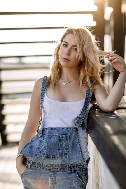 Young stylish blonde girl in denim overalls outdoors with natural daylight, a sunny day - Foto, immagini