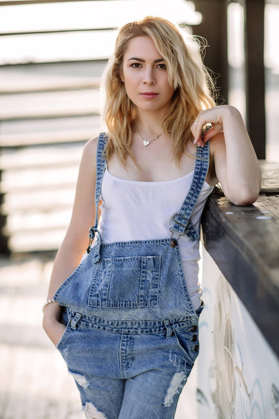 Young stylish blonde girl in denim overalls outdoors with natural daylight, a sunny day - Valokuva, kuva