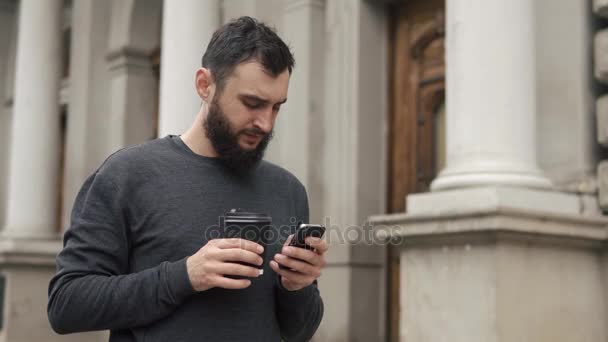 Happy beard young man texting on smartphone and drinking coffee in the street at sunset. He walks down the street, motion camera - Imágenes, Vídeo