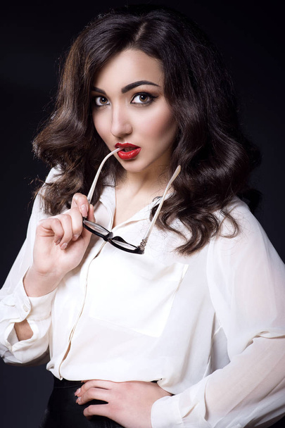 Kaunis nuori liiketoiminnan nainen tumma aaltoileva tukka ja punaiset huulet yllään valkoinen silkki pusero pitää puolella lasit suussaan
 - Valokuva, kuva