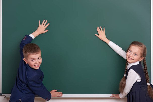 basisschool jongen en meisje handen zetten met schoolbord achtergrond en tonen van lege ruimte, gekleed in een klassieke zwarte pak, leerling van de groep, onderwijs concept - Foto, afbeelding
