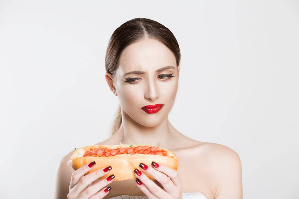 Unhealthy fast food. Beautiful young woman holding tasty hot-dog in her hands, thinking to eat it or not, having doubts about hotdog burger isolated white background. Face expression, body language - Photo, Image