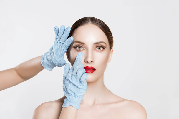 Beauty, people and health concept. beautiful young woman getting ready for eyelid lift, blepharoplasty plastic surgery doctors's hands in blue gloves pointing fingers to her eye over white background. - Photo, Image