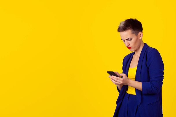 Portrait young angry woman unhappy, annoyed by something, someone on her cell phone texting, receiving bad sms text message news isolated yellow wall background. Human face expression emotion reaction - Foto, imagen