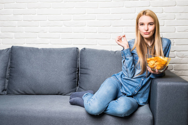 Молодая женщина смотрит телевизор и ест чипсы на диване
 - Фото, изображение