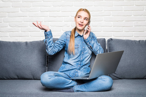 Smiling woman holding a smartphone and calling while using laptop on sofa - Photo, image