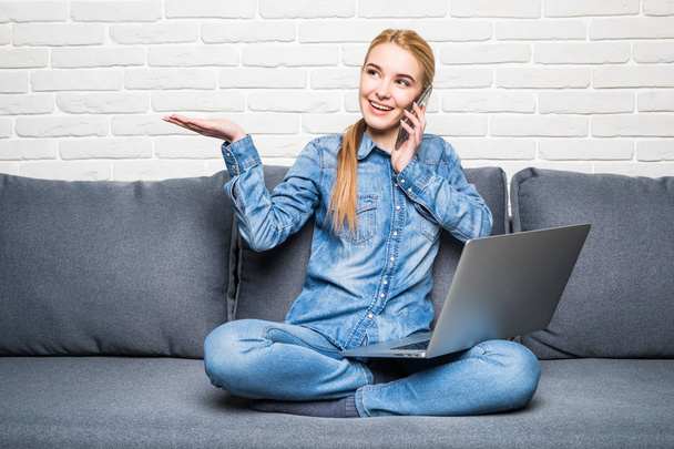 笑顔の女性がスマート フォンを押しながらソファーでノート パソコンを使用している間の呼び出し - 写真・画像
