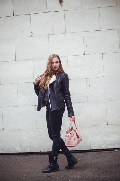 Portrait of a beautiful blonde young caucasian woman hipster In a black fur jacket posing fashion next to the white brick wall with pink bag. windy weather city portrait urban - Photo, Image