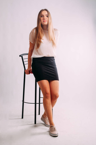 studio portrait of beautiful thin blonde caucasian young woman girl, isolated on white background posing in a white shirt and black office skirt. standing near red bar stool and doing fashion poses - Photo, Image