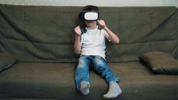 Little boy palting racing game using virtual reality headset sitting on the sofa - Кадри, відео