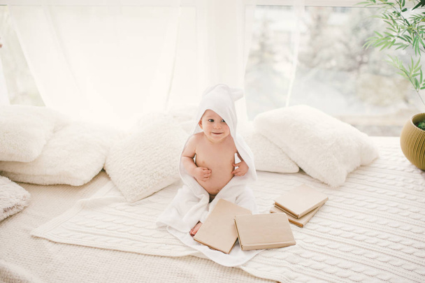 the kid in the white bamboo towel with ears sitting on a knitted blanket with books in the background of a large window. - Fotoğraf, Görsel