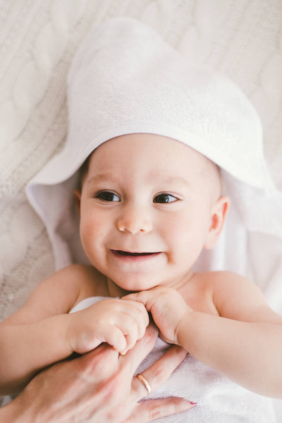 schöne lächelnde neugeborene Junge mit weißen Bambushandtuch mit lustigen Ohren bedeckt. liegt auf einem weiß gestrickten, wollkarierten hellen Interieur. Die Hand der Mutter liegt auf dem Kind. das natürliche Licht - Foto, Bild