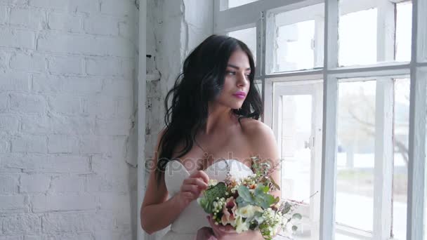 Bruid mooie brunette staande in bruiloft jurk met boeket in de buurt van venster - Video