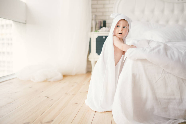 новорожденный 5-месячный ребенок в спальне рядом с большой белой кроватью на деревянном полу, завернутый в белое бамбуковое полотенце
. - Фото, изображение