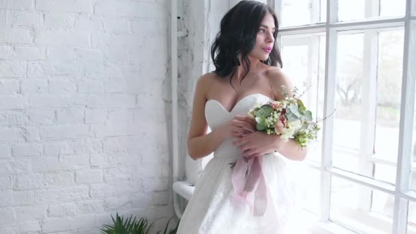 Nevěsta, krásná bruneta postavení v svatební šaty s kytici okna - Záběry, video