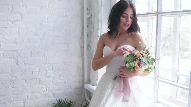 Braut schöne Brünette steht im Brautkleid mit Blumenstrauß am Fenster - Filmmaterial, Video