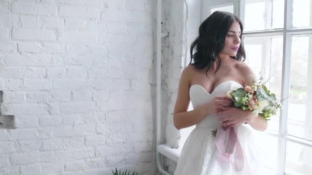 Bruid mooie brunette staande in bruiloft jurk met boeket in de buurt van venster - Video