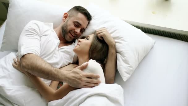 Young beautiful and loving couple talk and hug into bed while waking up in the morning. Top view of attractive man chatting his smiling wife - Záběry, video