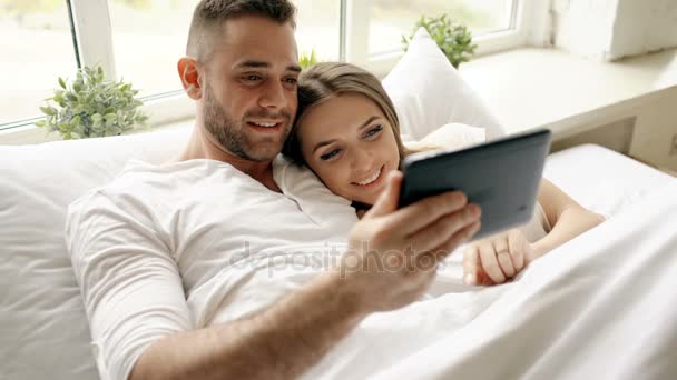 Young cute and loving couple using tablet computer and talking lying in bed in the bedroom at morning - Materiał filmowy, wideo