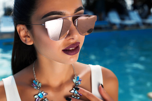 Summer fashion close up portrait of beautiful sexy woman in white stylish swimwear and mirrored sunglasses with perfect necklace and  hairstyle at swimming pool background - Valokuva, kuva