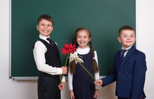 niños dando flores de niña, niño de la escuela primaria cerca de fondo de pizarra en blanco, vestido con traje negro clásico, alumno de grupo, concepto de educación
 - Foto, Imagen