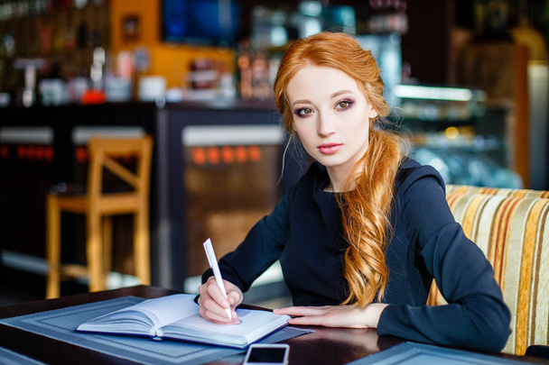Thoughtful young businesswoman sitting in a cafe and writing in notepad - Zdjęcie, obraz