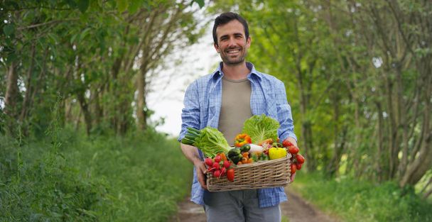 Portrait of a happy young farmer holding fresh vegetables in a basket. On a background of nature The concept of biological, bio products, bio ecology, grown by own hands, vegetarians, salads healthy - Foto, imagen