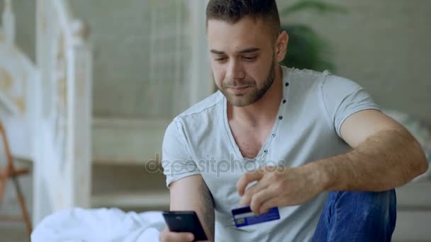 Attractive young man with smartphone and credit card shopping on the internet sit on bed at home - Metraje, vídeo