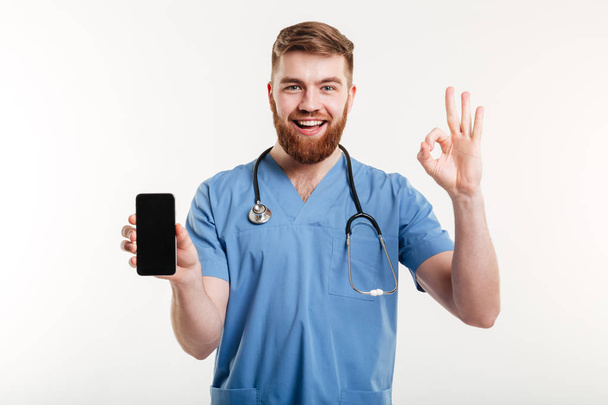 Male doctor smiling with phone and showing gesture - Foto, immagini