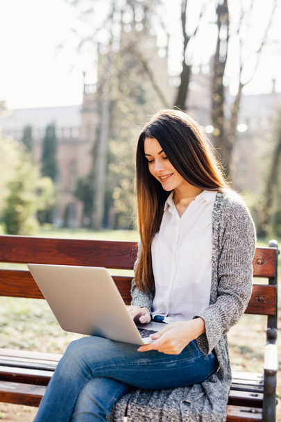 πορτρέτο μιας νεαρής γυναίκας επαγγελματική χρησιμοποιώντας ένα φορητό υπολογιστή, ενώ κάθεται σε ένα παγκάκι σε ένα πάρκο, σε μια ηλιόλουστη ημέρα. - Φωτογραφία, εικόνα