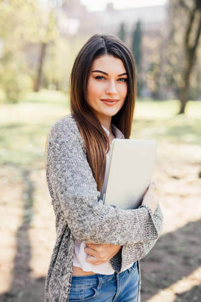 Mooie jonge vrouw lopen met een laptop onder de hand in park - Foto, afbeelding
