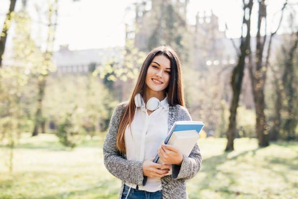 fröhliche attraktive junge Frau mit Notizbüchern, die im Park steht und lächelt - Foto, Bild