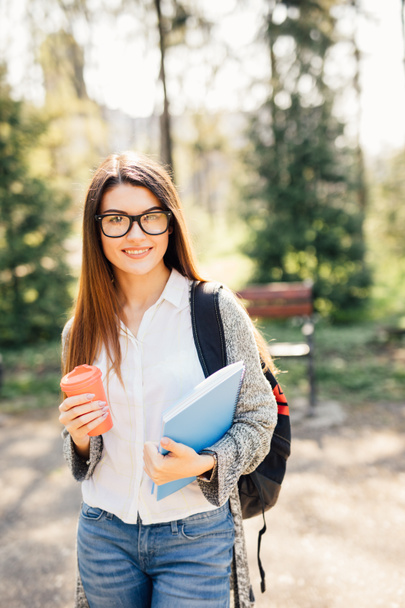 Female student outdoors holding a notebook and smiling with cup of coffee - Photo, image