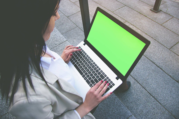 Useful gadget for business. Low angle closeup view of beautiful business woman in smart casual wear working on laptop while sitting outdoors. Green chroma key screen on laptop. - 写真・画像
