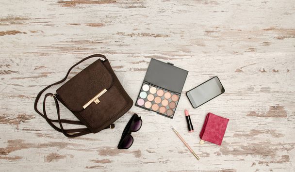 Fashionable concept: eyeshadows, handbag, glasses, mobile phone, lipstick, wallet on a wooden background. top view - Фото, изображение