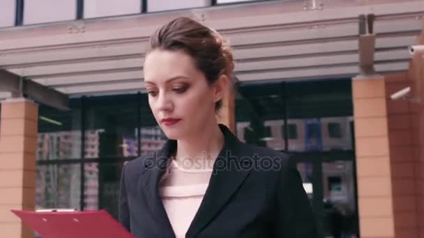 Beautiful confident young business woman is in a hurry to negotiate and looks at the clock. Girl in business clothes with a red folder in hands on the background of an office building - Séquence, vidéo