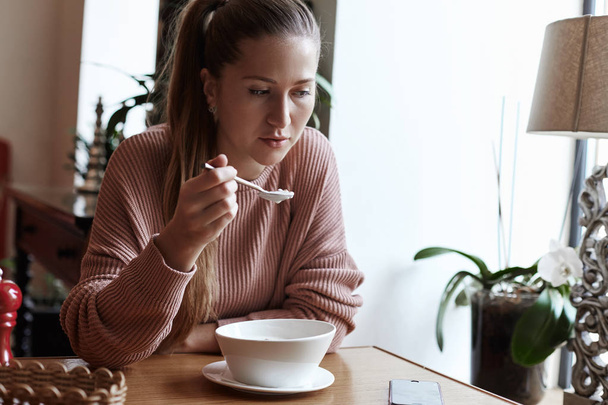 Healthy eating, balanced diet, food and people concept. Young Caucasian attractive lady is having her breakfast bowl with granola and farm yogurt in organic modern cafe. Beautiful girl is looking at her phone, waiting for message while her eating . - Foto, imagen