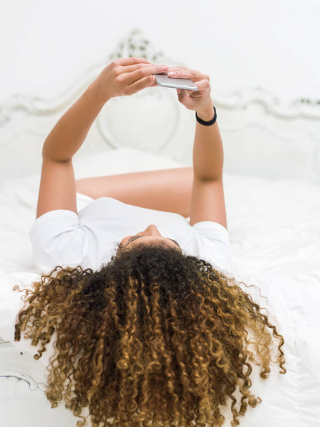 Retrato de mujer con hermoso cabello acostado en la cama y tomando selfie
 - Foto, imagen