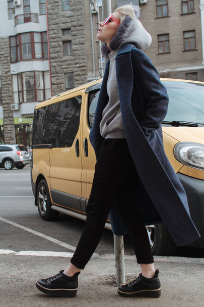 Μια ξανθιά κοπέλα με τα γυαλιά και ένα παλτό βόλτες στους δρόμους της πόλης - Φωτογραφία, εικόνα