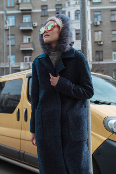 Egy szőke lány, egy kabátot és szemüveg sétálni a város utcáin - Fotó, kép