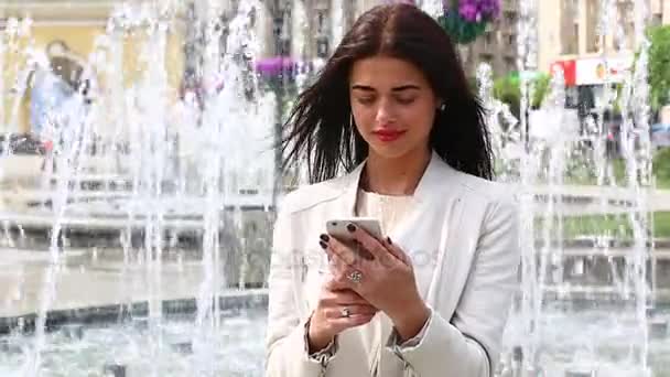 Portrait de fille riant à l'aide d'un smartphone. Jolie jeune femme avec son téléphone portable sur fond de fontaine. Jolie femme heureuse utilisant un smartphone dans le parc de la ville, Steadicam shot
. - Séquence, vidéo