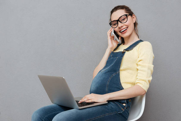 Pregnant happy lady using laptop computer and talking by phone. - Foto, Imagen