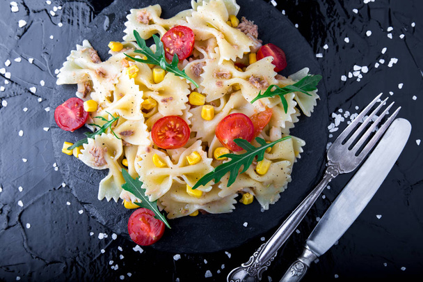 スレート板トマト チェリー、ツナ、コーン、ナイフやスプーンの近くルッコラのパスタサラダ。平面図です。食材。イタリア料理. - 写真・画像