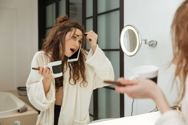 Kobieta robi makijaż i rozmowy na telefon komórkowy w łazience - Zdjęcie, obraz