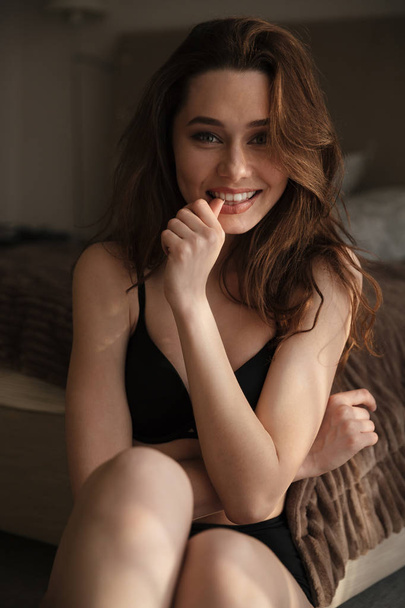 Cheerful beautiful young woman in lingerie sitting in bedroom - 写真・画像