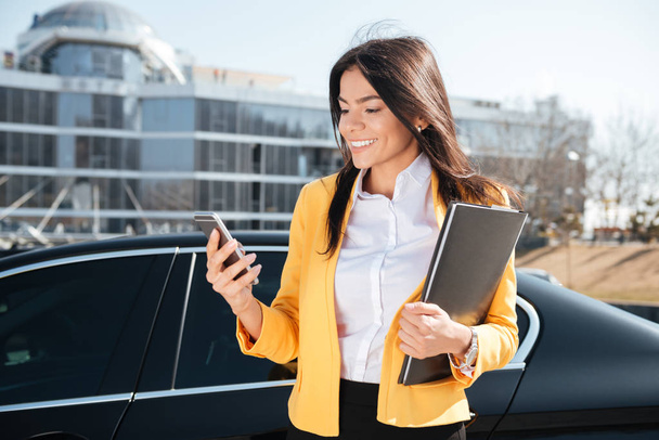 Smiling young businesswoman with documents in folder using smartphone outdoors - Foto, imagen