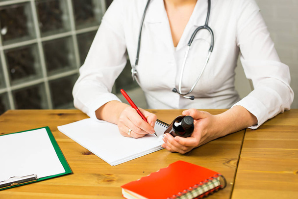 Close-up of female physician medicine doctor or pharmacist sitting at work table, holding jar or bottle of pills in hand and writing prescription on special form. Medical care, pharmacy or health - Φωτογραφία, εικόνα